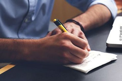 Ein Mann h&auml;lt einen Kugelschreiber in seiner rechten Hand und macht sich Anmerkungen auf einem kleinen Notizblock.