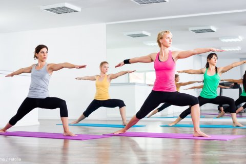 Frauen stehen in einer aufrechten Yoga-Position mit einem Ausfallschritt und ausgestreckten Armen auf Yogamatten in einem Kursraum mit einem Spiel im Hintergrund