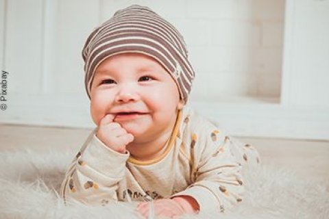 Lächelndes Baby liegt auf dem Bauch eines Teppichs mit erhobenem Kopf und einem Finger im Mund