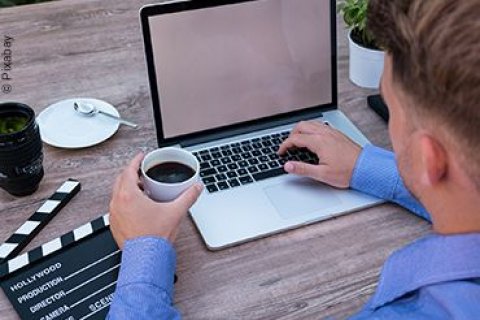 Ein Business-Mann sitzt vor seinem Laptop mit einem Kaffee in der einen Hand und mit der anderen Hand tippend auf der Tastatur des Laptops.