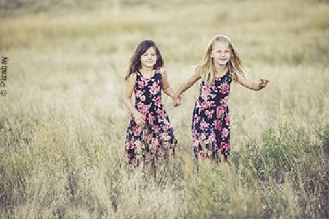 Zwei Mädchen laufen Händchenhaltend im Sommerkleid durch ein Kornfeld.
